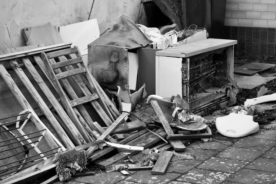 An outdoor scene showing a pile of discarded household items and debris on a paved pavement outside a building, with a brick wall visible in the background. The debris includes broken wooden furniture, such as a disassembled wooden chair and a ladder, along with pieces of torn fabric, plastic wrapping, cardboard, and miscellaneous loose materials. There is a partially open cabinet with exposed damaged internal components, and some damaged appliances or containers among the rubbish. A white plastic item, possibly a part of an appliance or protective packaging, is lying on the ground. The scene appears to depict a collection of waste or bulky items awaiting removal, with some items stacked and others scattered, suggesting the process of clearing or preparing for house removals. The setting is lit with natural light, highlighting the disorder and materials involved in home relocation or waste disposal, as handled by service providers like Man and a Van Marylebone.