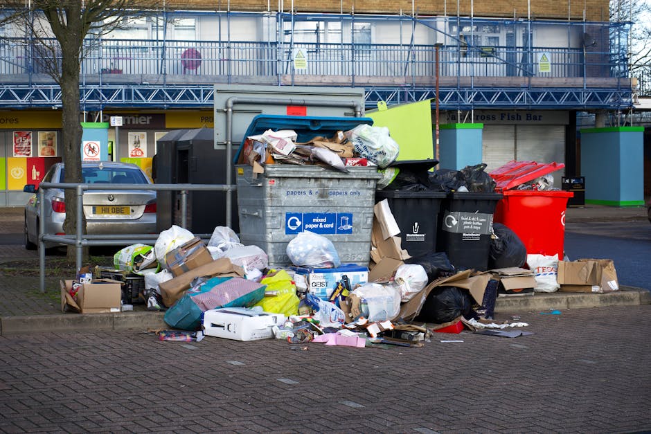An outdoor scene showing a pile of discarded household items and debris on a paved pavement outside a building, with a brick wall visible in the background. The debris includes broken wooden furniture, such as a disassembled wooden chair and a ladder, along with pieces of torn fabric, plastic wrapping, cardboard, and miscellaneous loose materials. There is a partially open cabinet with exposed damaged internal components, and some damaged appliances or containers among the rubbish. A white plastic item, possibly a part of an appliance or protective packaging, is lying on the ground. The scene appears to depict a collection of waste or bulky items awaiting removal, with some items stacked and others scattered, suggesting the process of clearing or preparing for house removals. The setting is lit with natural light, highlighting the disorder and materials involved in home relocation or waste disposal, as handled by service providers like Man and a Van Marylebone.