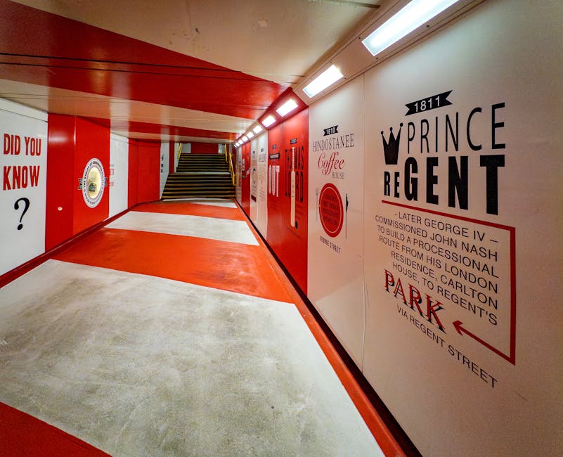 A brightly lit underground corridor featuring red and white painted walls with vintage-style signage and advertisements, including a large sign for 'Prince Regent' from 1811, along with text about George IV and historical building information. The floor is concrete with a red-painted section leading to a staircase with metal handrails, situated at the end of the corridor. The ceiling has a combination of white panels and exposed lighting fixtures. On the right side, the corridor opens to an area where furniture and packing materials, such as cardboard boxes wrapped in plastic and fabric blankets, are being loaded onto a large moving van parked outside. A person in casual clothing is visible, actively lifting and carrying a cardboard box, coordinating logistics for a home relocation or furniture transport. The scene aligns with house removals or moving services, highlighting the packing, loading process, and interior environment typical of professional removals as managed by [COMPANY_NAME].