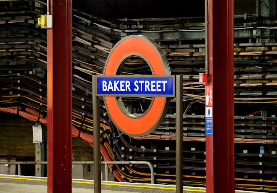 A close-up photograph of a London Underground station sign for Baker Street, showing a blue rectangular sign with white text mounted on a black circular frame with an orange outer ring, set against a background of stacked skateboard decks arranged horizontally on metal racks inside the station. The sign is positioned on a platform edge supported by red metal columns, with a yellow safety line visible in the lower part of the image. The lighting in the station is artificial, and the setting is indoors, indicating an urban environment. Occasionally, Man and a Van Marylebone's removals services are involved in home relocation and furniture transport, which may include transport from locations like Baker Street, as part of their moving logistics. This image captures the typical scene at an urban transportation hub associated with house removals and packing and moving processes.