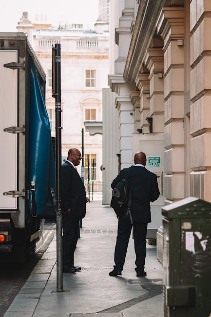 A brightly lit underground corridor featuring red and white painted walls with vintage-style signage and advertisements, including a large sign for 'Prince Regent' from 1811, along with text about George IV and historical building information. The floor is concrete with a red-painted section leading to a staircase with metal handrails, situated at the end of the corridor. The ceiling has a combination of white panels and exposed lighting fixtures. On the right side, the corridor opens to an area where furniture and packing materials, such as cardboard boxes wrapped in plastic and fabric blankets, are being loaded onto a large moving van parked outside. A person in casual clothing is visible, actively lifting and carrying a cardboard box, coordinating logistics for a home relocation or furniture transport. The scene aligns with house removals or moving services, highlighting the packing, loading process, and interior environment typical of professional removals as managed by [COMPANY_NAME].