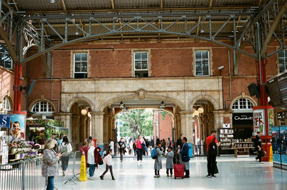 Photo of a busy station interior with a high, arched ceiling supported by metal beams and iron framework, featuring exposed brickwork on the upper walls. The large entrance opening at the center reveals a view of green trees outside. Inside, there are several people, including adults and children, some holding bags or suitcases, indicating a home relocation or travel setting. On the left side, a flower stand with various plants and flowers is visible, while on the right, a food or gift shop displays shelves and signage, including a sign for 'Hotel Chocolate.' Individuals are walking, waiting, and engaging in activities related to packing and moving logistics, typical of a busy transport hub. The scene captures the movement of luggage and the indoor environment suited for furniture transport and associated moving services, with lighting that combines natural daylight from outside and interior fixtures.