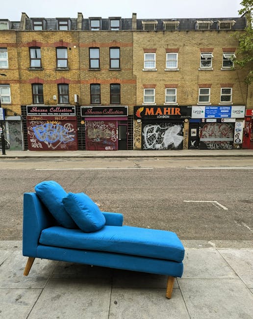 A blue fabric upholstered chaise lounge with matching cushions positioned on a concrete sidewalk beside a street in Marylebone, London. The furniture is set directly outside a row of closed shopfronts, some of which are covered in colorful graffiti and signage, including a shop named 'Mahir' and other storefronts with roller shutters. The chaise lounge is oriented horizontally across the lower part of the image, with its backrest and seat cushions visible, and is prepared for removal as part of a home relocation or furniture transport process. The scene is under natural daylight, with a brick building in the background featuring multiple windows and a flat roof. This image may be used to illustrate furniture disposal or a removal company's operational context in urban areas, such as Man and a Van Marylebone's services for bulky-item challenges like sofa disposal.