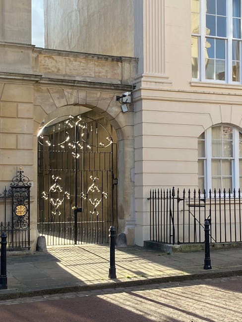 Photograph of a historic building entrance in Marylebone during daylight, with a large wrought iron gate set within a stone archway, part of a beige sandstone facade featuring classical architectural details. The gate is partially open, revealing the interior courtyard illuminated by sunlight, which casts shadow patterns onto the pavement. To the left of the gate, there is a decorative black iron fence with ornate scrollwork and a small gate, and to the right, a black metal railing with vertical bars. The building has tall windows with white frames, large glass panes, and decorative molding along the exterior wall. The scene suggests a setting suitable for home relocation or furniture transport, with the open gate indicating access for moving or loading items, and a hint of the building's historic character associated with professional removals services such as those offered by Man and a Van Marylebone.