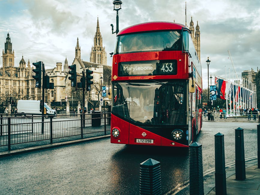 A red double-decker bus with route number 453 displaying 'Marylebone' on its front, is stopped on a wet urban street surface near a platform with black bollards, in front of historic Gothic-style buil
