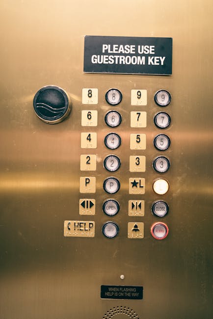 Close-up view of an elevator control panel inside a building used for house removals and moving logistics. The panel features a black sign at the top that reads 'PLEASE USE GUESTROOM KEY', with a large black circular button to its left. Below, there are numbered buttons from 1 to 9 and 0, arranged in a grid, alongside floor indicators with small circular displays; some rooms are marked with white labels such as 'P' for parking and an 'L' with a star symbol. Additional buttons include 'OPEN', 'CLOSE', and 'HELP', with symbols representing door operation. The panel also has a red emergency stop button and a note at the bottom stating 'WHEN FLASHING HELP IS ON THE WAY.' The setting appears to be in a commercial or residential building likely involved in home relocation services, with the control panel designed for managing access during moves or relocations.