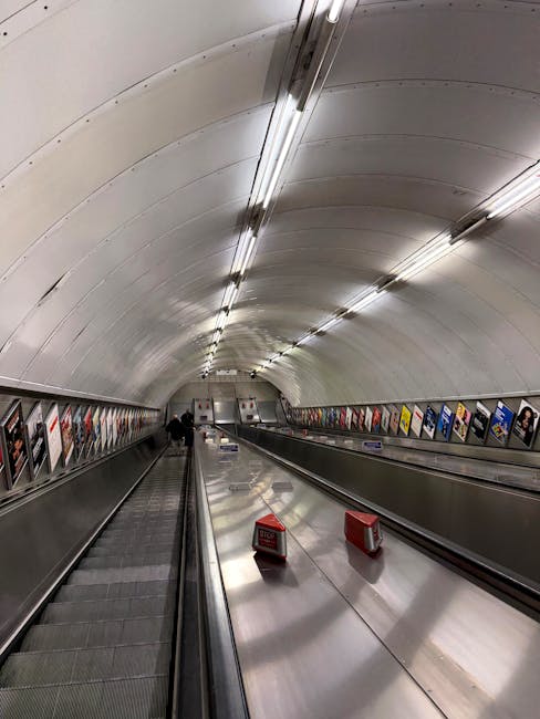 An underground pedestrian tunnel with a curved, white ceiling fitted with evenly spaced fluorescent lighting. The tunnel has two metallic moving walkways separated by a central strip, with one walkway visible on the left and an empty escalator on the right. Along the sides, there are advertising posters in brightly coloured frames attached to the walls. Near the foreground, two red warning signs are placed on the moving walkway, indicating caution. The environment appears clean and modern, typical of a public transport connection point used during house removals for navigating between stations or access points, supported by the professional services of Man and a Van Marylebone.