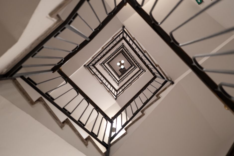 A multi-story staircase viewed from above, showing a series of square landings and metal railings painted black, inside a building with white walls. The staircase is enclosed within the interior space, and a central light fixture at the top provides illumination. The image captures the vertical layout and construction of the staircase, illustrating potential challenges in moving furniture or packing boxes between floors during a home relocation or removal service. Man and a Van Marylebone specializes in furniture transport and packing for house removals, often managing access issues like staircase and lift difficulties, as seen in this environment. The photo highlights the internal environment where careful planning is necessary to navigate narrow or complex staircases during the loading process, emphasizing the importance of professional removals services for efficient and safe relocation solutions.