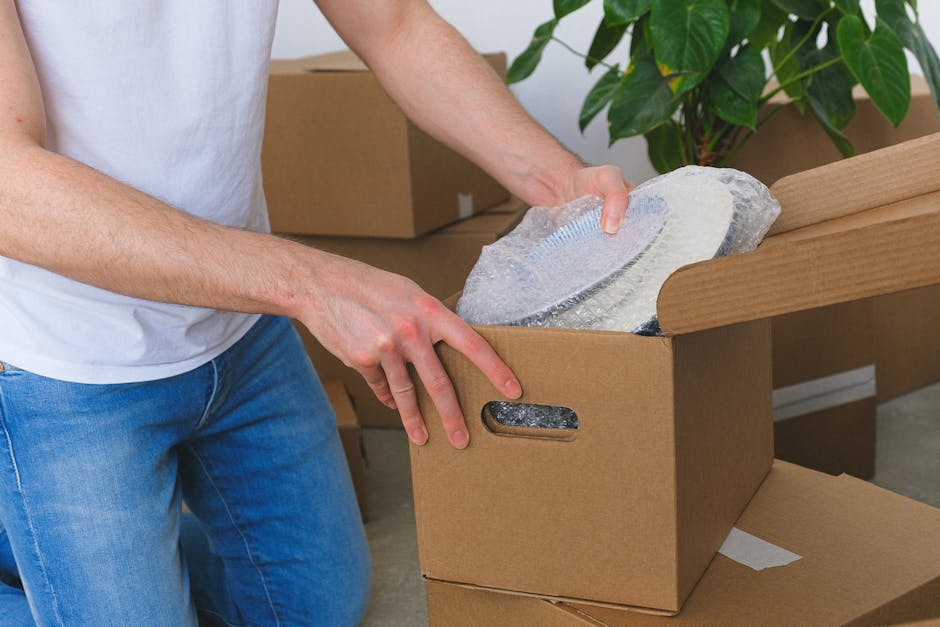 A person dressed in a white t-shirt and blue jeans is in the process of preparing a cardboard box for a home relocation, as part of a furniture transport or packing and moving activity. The individual is seen holding a protective wrap or bubble wrap and placing it over a fragile item, possibly a dish or glassware, which is partially covered in additional packing material for safety during transit. The scene takes place inside a property, with other packing supplies nearby, including additional cardboard boxes stacked in the background. The room has natural lighting, illuminating the wooden surface of the box and the greenery of a potted plant nearby. This moment captures the careful packing process, emphasizing proper handling techniques to prevent damage during removals, and reflects the logistical considerations involved in professional house removals, as executed by companies like Man and a Van Marylebone.