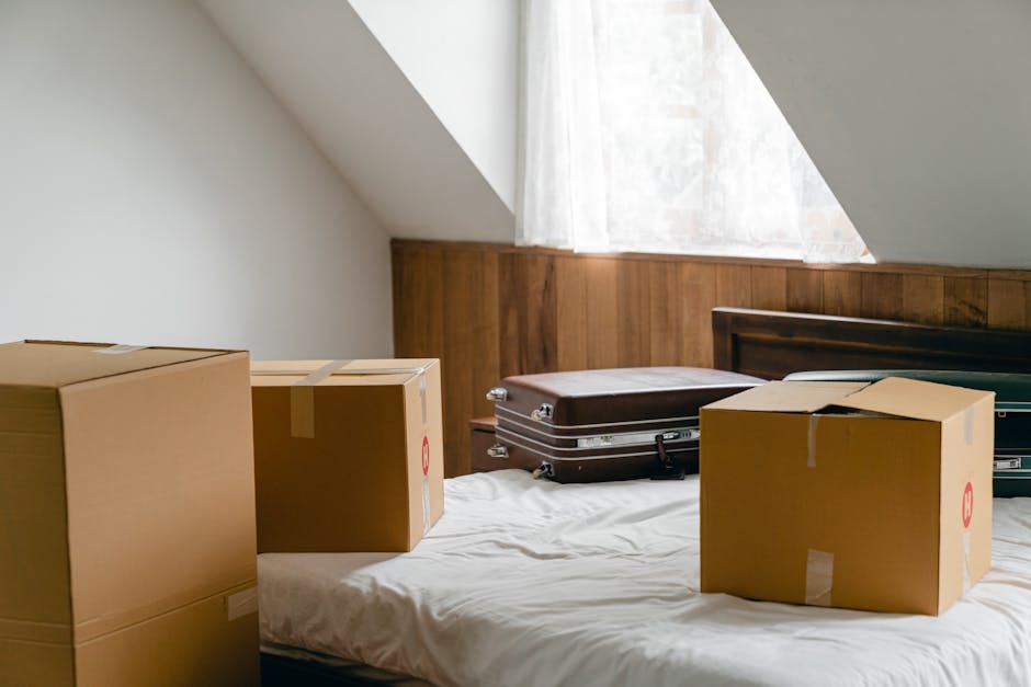A person dressed in a white t-shirt and blue jeans is in the process of preparing a cardboard box for a home relocation, as part of a furniture transport or packing and moving activity. The individual is seen holding a protective wrap or bubble wrap and placing it over a fragile item, possibly a dish or glassware, which is partially covered in additional packing material for safety during transit. The scene takes place inside a property, with other packing supplies nearby, including additional cardboard boxes stacked in the background. The room has natural lighting, illuminating the wooden surface of the box and the greenery of a potted plant nearby. This moment captures the careful packing process, emphasizing proper handling techniques to prevent damage during removals, and reflects the logistical considerations involved in professional house removals, as executed by companies like Man and a Van Marylebone.