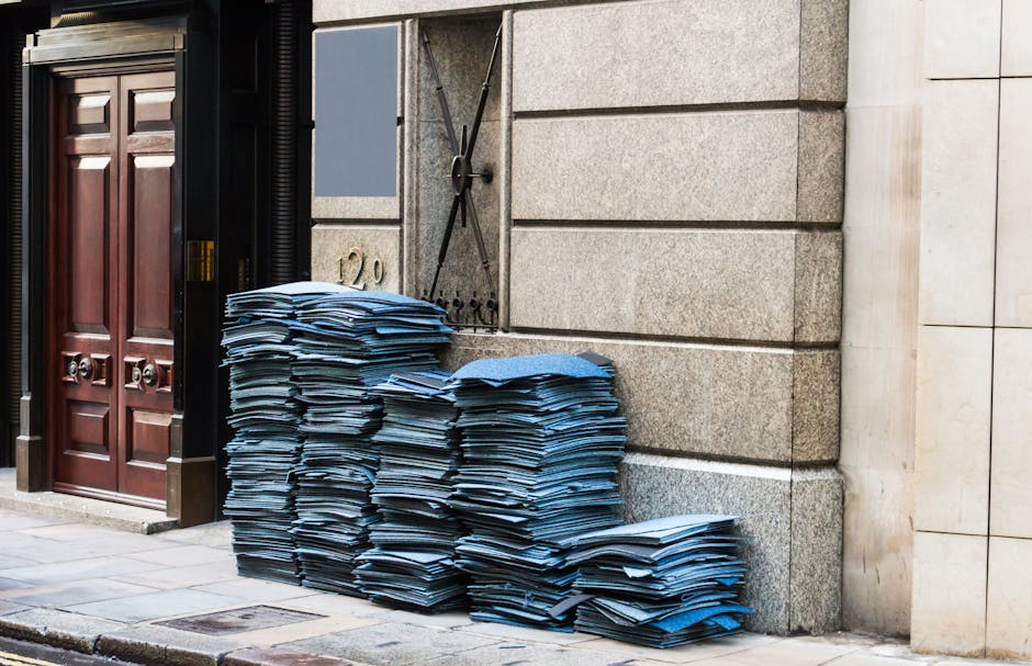 Stacks of blue and black cardboard folders and packaging materials piled on the pavement outside a building’s entrance, adjacent to a dark wooden door and a gray stone wall with a decorative black metal gate. The folders are arranged in multiple neat piles, some leaning slightly, with a few scattered at the base. The scene is illuminated by natural daylight, and the setting appears to be in a busy urban area, possibly during a home relocation or moving process. The image captures the external environment near the entryway, emphasizing the preparation phase of packing and loading for a house removal service, as provided by Man and a Van Marylebone, supporting efficient transportation and logistics in accordance with Westminster Council kerbside loading permits.