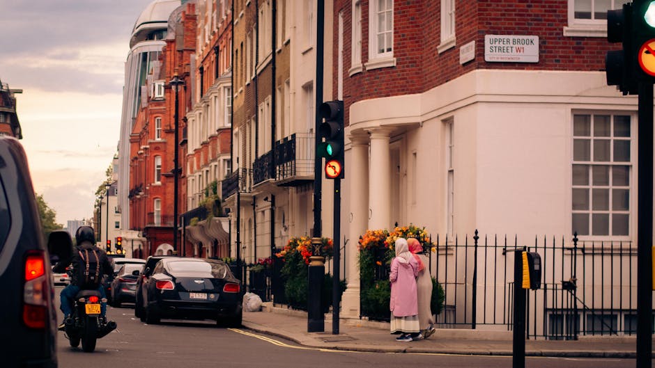 A street scene on Upper Brook Street in W1, Marylebone, showing a row of Georgian-style terraced houses with red brick facades and white window frames. In the foreground, a black leather armchair, a cardboard box, and a plastic-wrapped wrapped mirror are positioned on the pavement, likely prepared for a home relocation. A man on a motorcycle and several parked cars are visible along the street. A woman dressed in a pink coat and headscarf, accompanied by a young girl, are standing near a black wrought iron fence outside one of the houses, possibly waiting or observing the loading process. Traffic lights and street signs are mounted on black posts, with flowering plants in large pots adding colour to the scene. The environment is illuminated by soft, natural evening light, capturing the ongoing house removals and transport preparations, in line with services provided by Man and a Van Marylebone for relocation logistics, packing, and furniture transport.