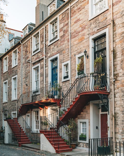 Exterior view of a multi-storey residential building with a stone façade, featuring a prominent red external staircase connecting the ground floor to the first-floor entrance, with black metal railings on both sides of the staircase. Potted plants and greenery are placed along the staircase and near the doorways, which vary in color including blue and red. A street scene shows the building situated on a cobblestone pavement, with neighboring buildings visible in the background. This setting captures the typical architecture of Marylebone, and the image reflects a property suitable for home relocation or furniture transport, possibly during a house removal process. [COMPANY_NAME], experienced in removals, often assists clients with such activities, including packing and loading in urban environments like this. Natural lighting highlights the textures of the stone façade and details of the staircase, providing a clear view of the building's exterior and access points for moving services.
